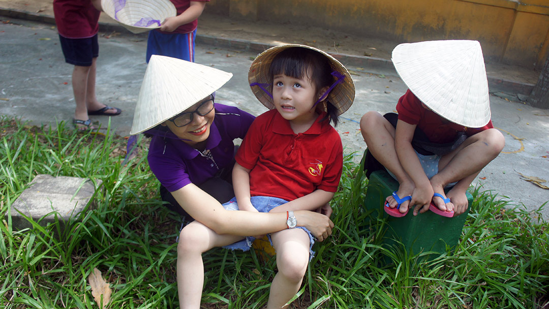 A girl in a Vietnamese hat sits on the grass with her teacher.