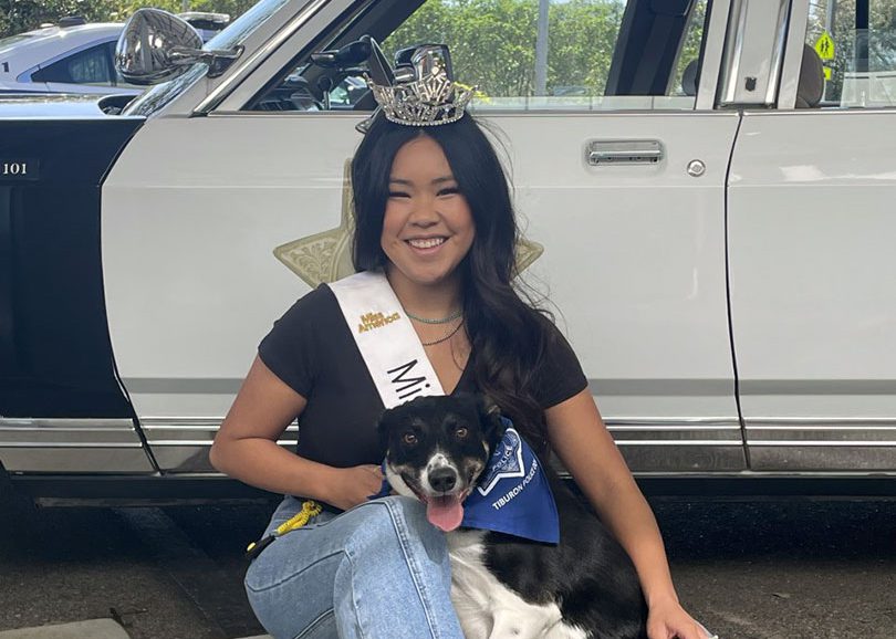 A woman in a tierra and sash sitting in front of a car with her dog