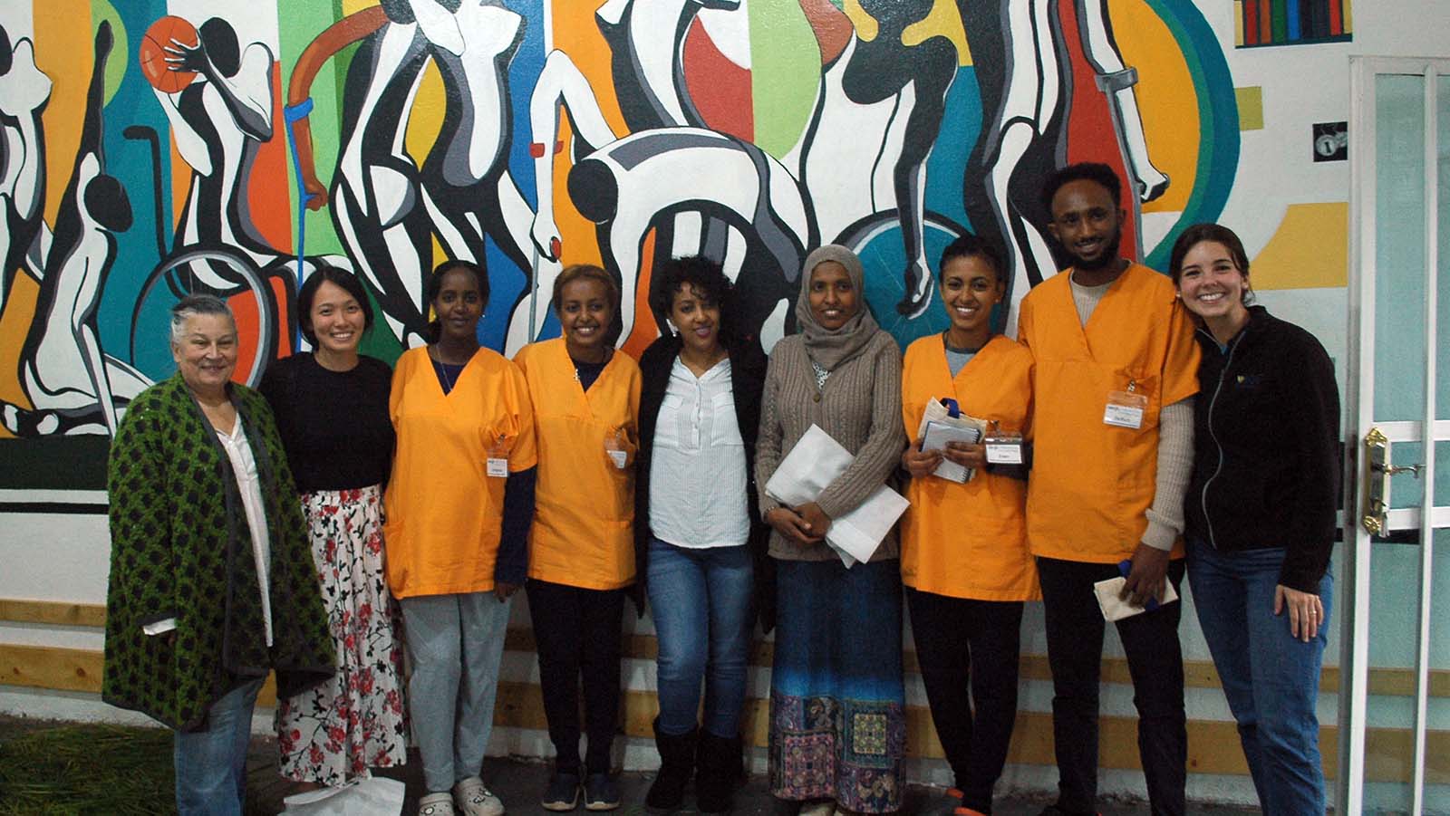 Group of smiling people stands in front of a colorful mural