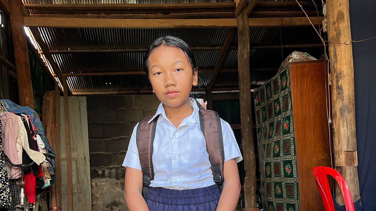 Girl in school uniform and backpack smiles in front of house in Cambodia