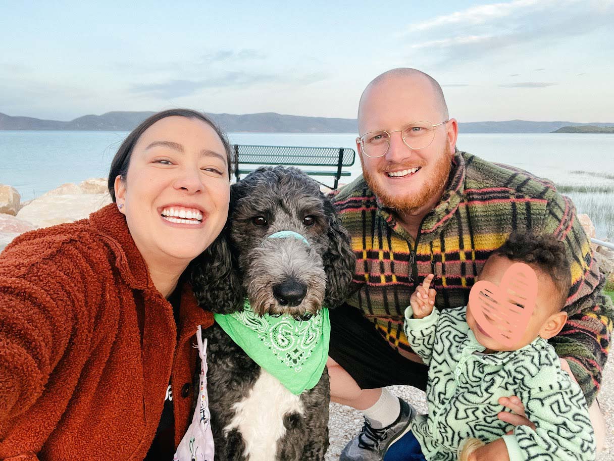 Family with toddler and dog takes a family picture on the shore of a lake
