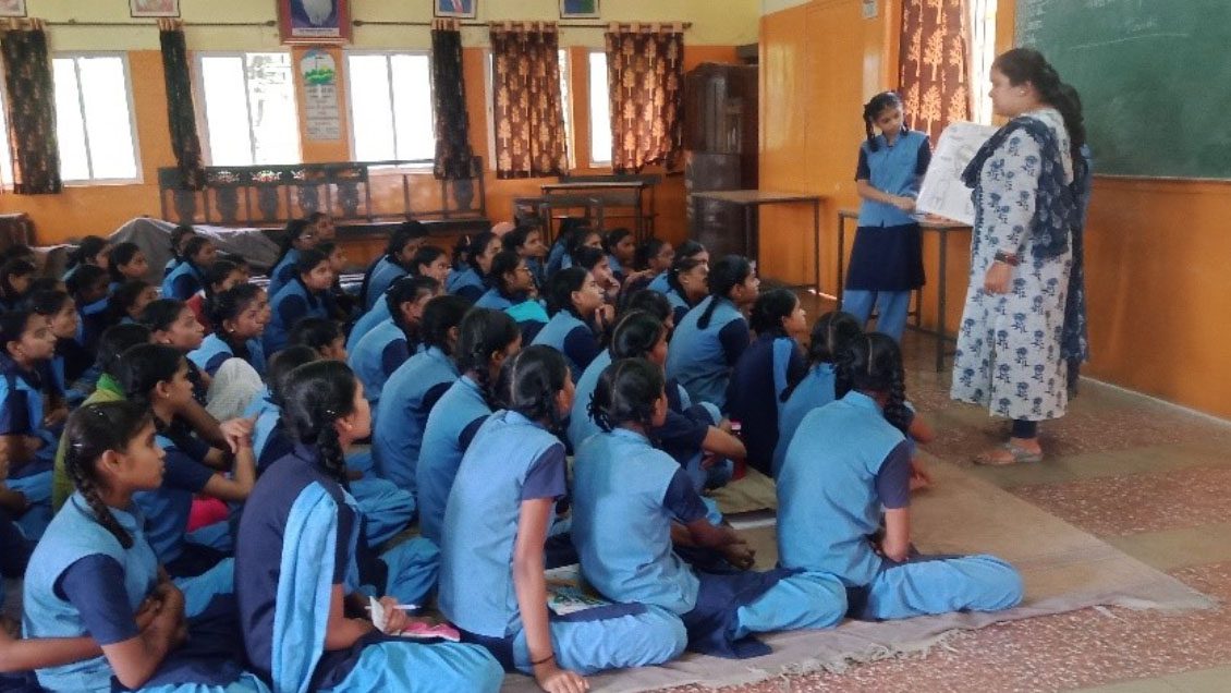 Class of girls in blue school uniforms look up at teacher by blackboard