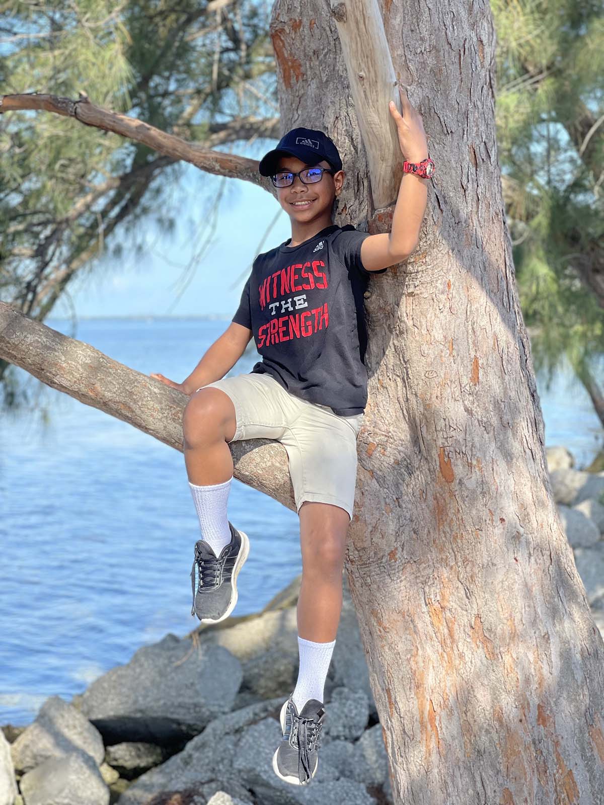 Boy sits on tree branch in front of lake and smiles