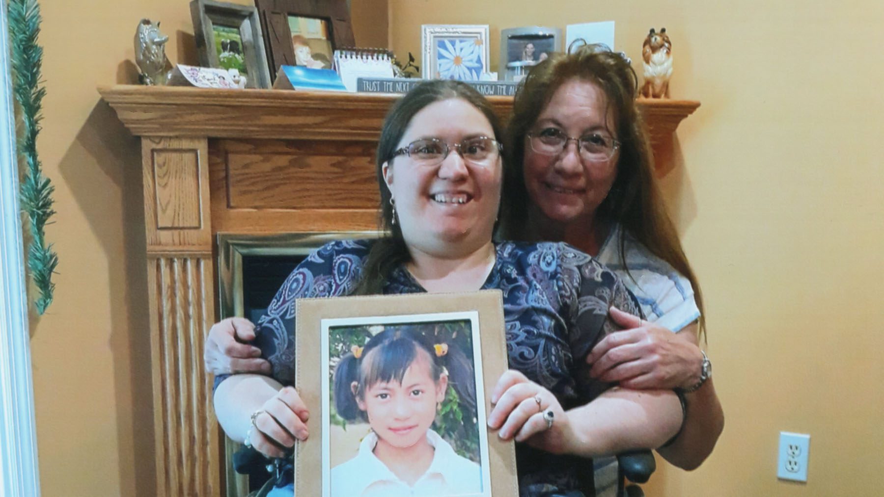 Mother and daughter in wheelchair stand in front of fireplace and hold framed picture of sponsored child