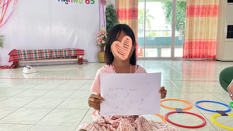 Girl holds up drawing while sitting on floor with other toys