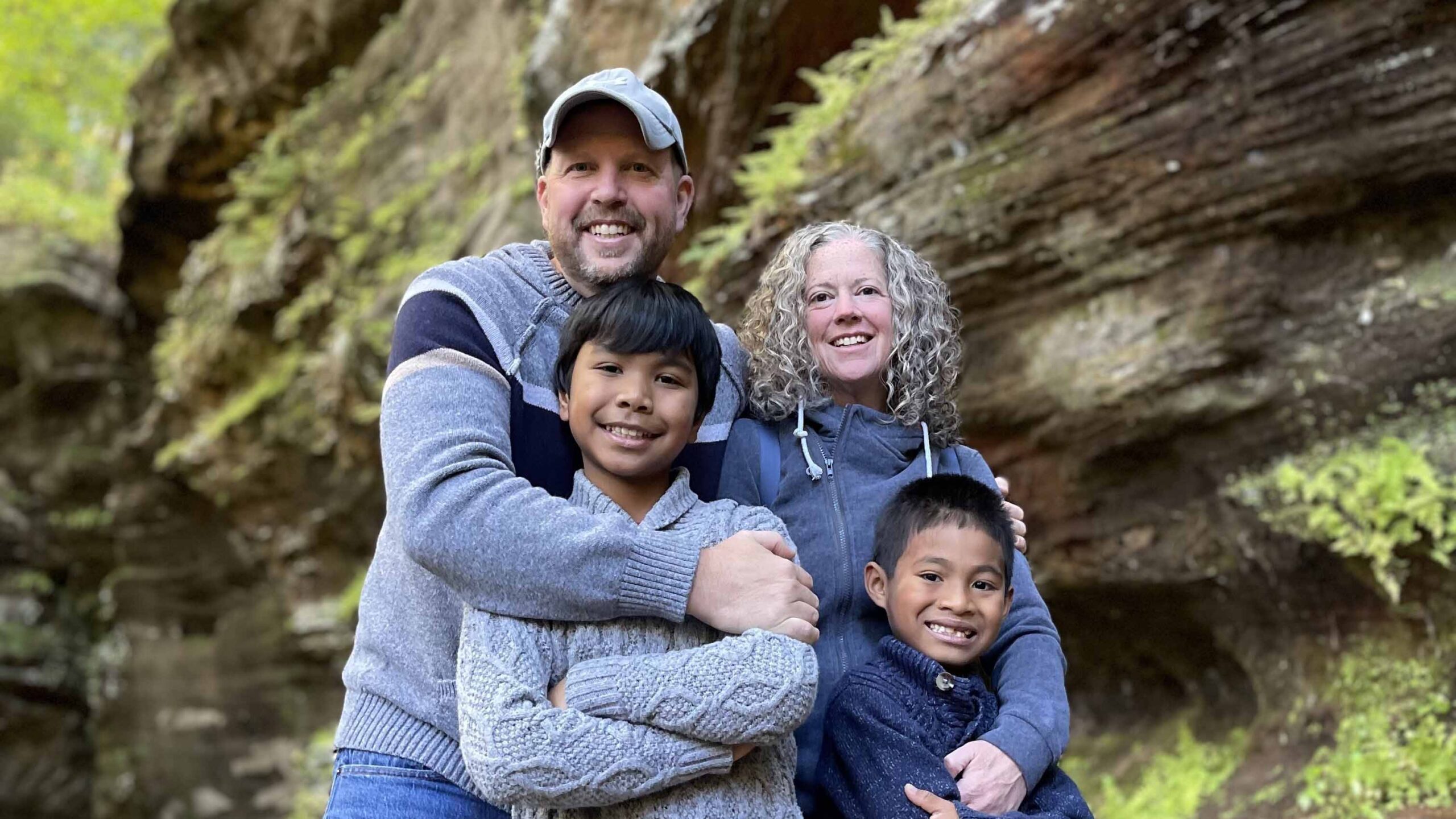 Family of four stands together outside in front of a mossy rock