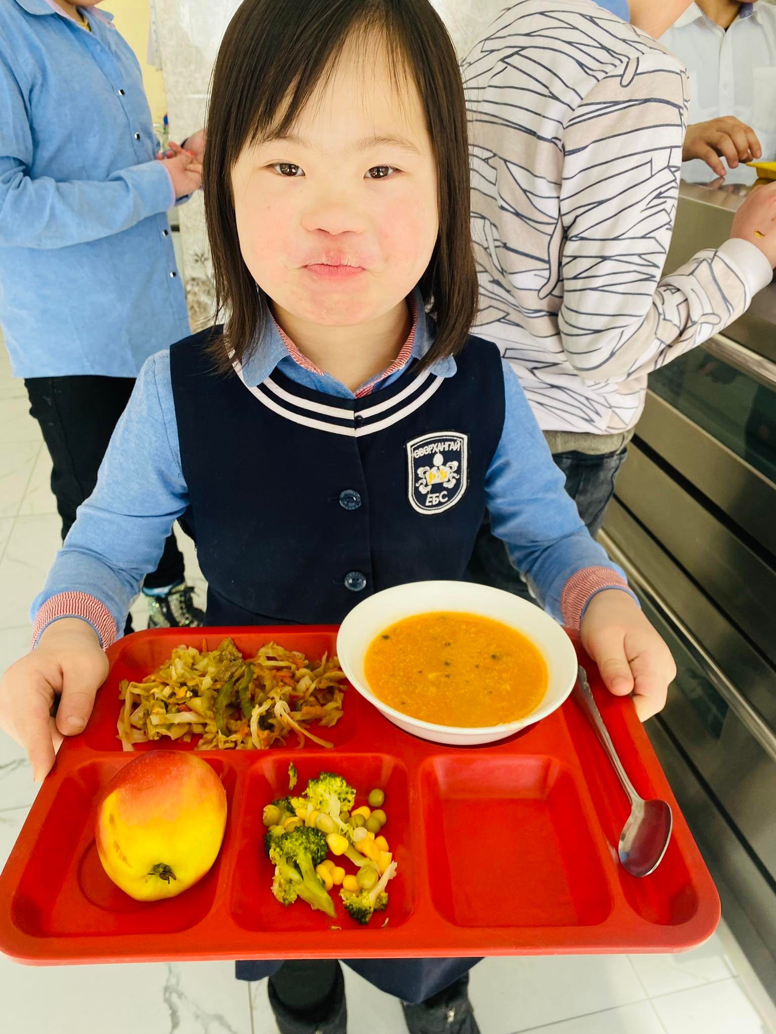 Little girl in school uniform smiles with school lunch.