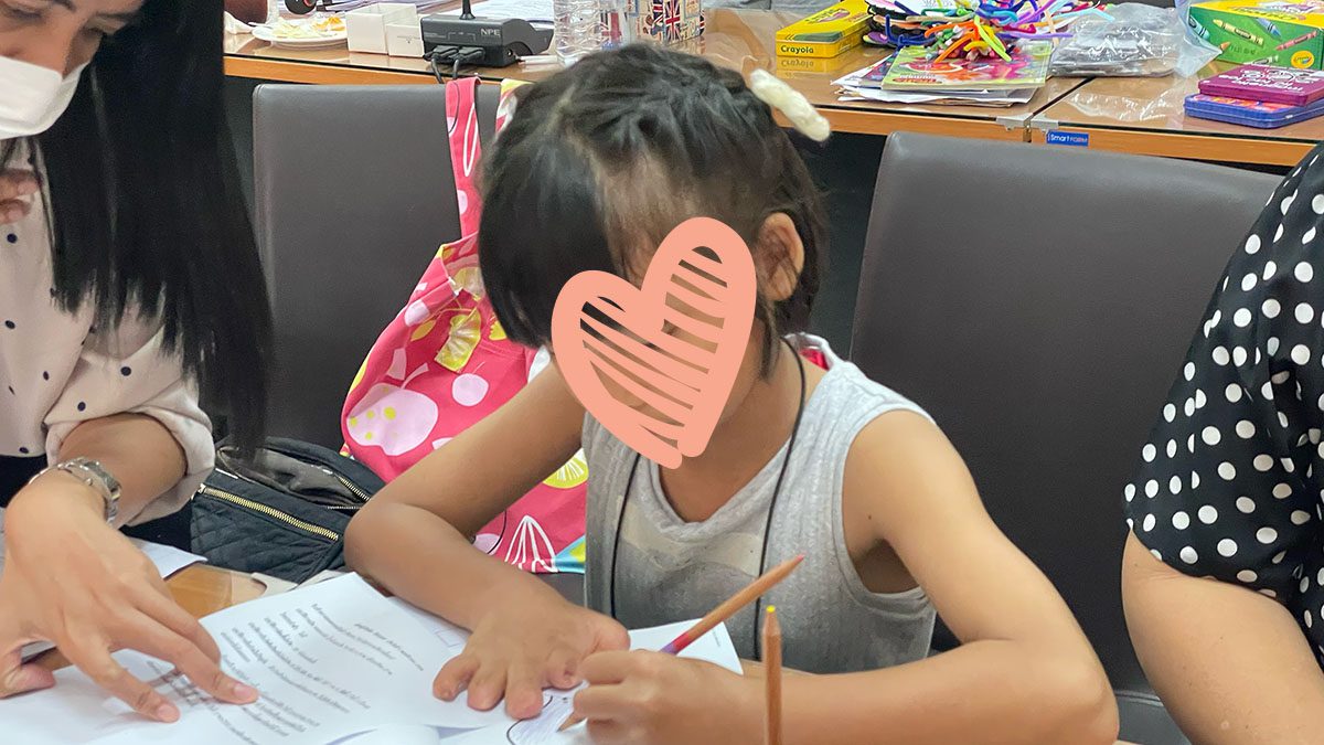 Little girl in a tank top sits at a table and writes with a pencil.