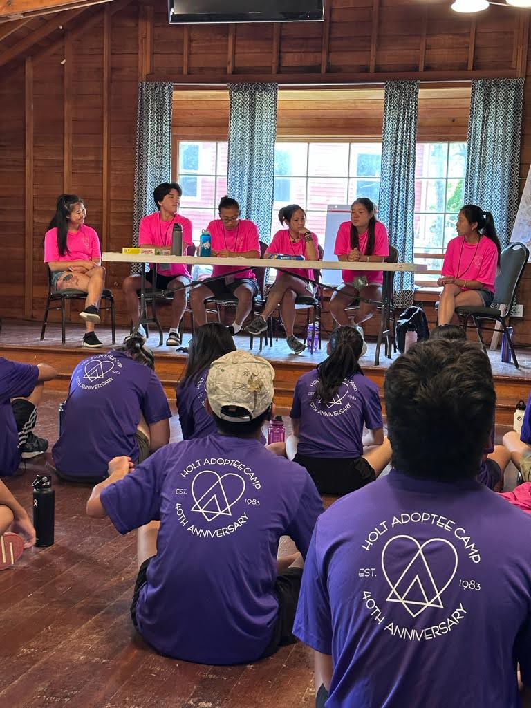 Panel of camp counselors sits around table while campers watch