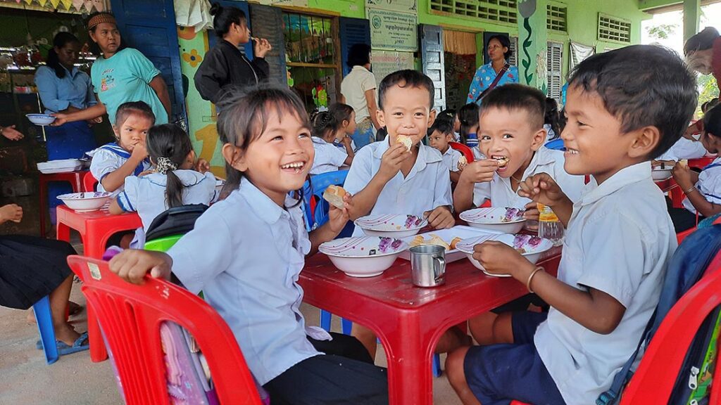 Preschoolers sit on red plastic chairs laughing as they eat a snack