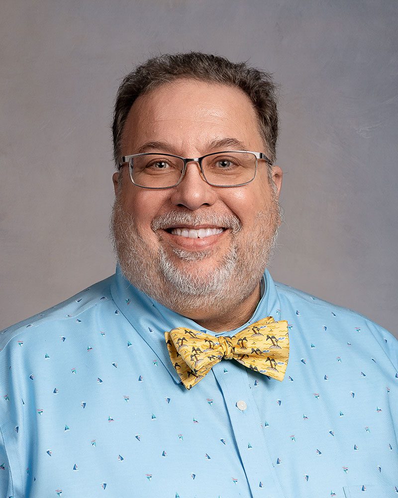 Headshot of a man in a blue shirt and yellow bowtie, wearing glasses