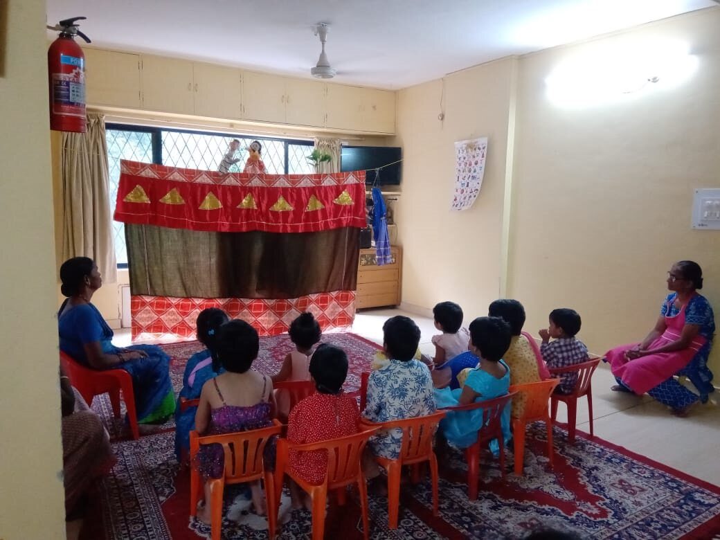 A group of children sits in small plastic chairs to watch a puppet show