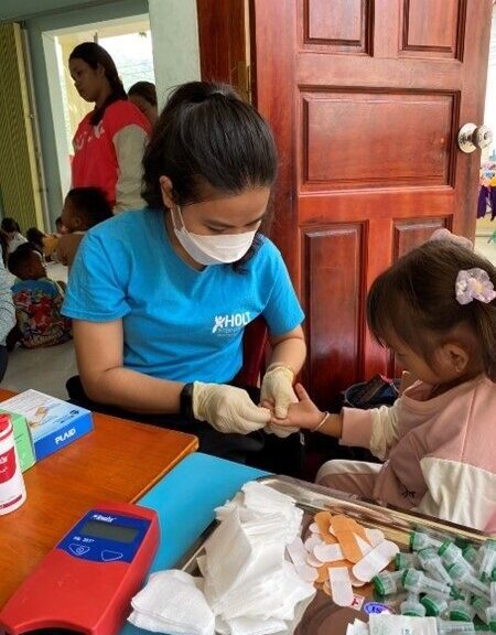 Healthcare worker in a mask screens a little girl in a pink shirt, next to a table with gauze, bandaids and tubes