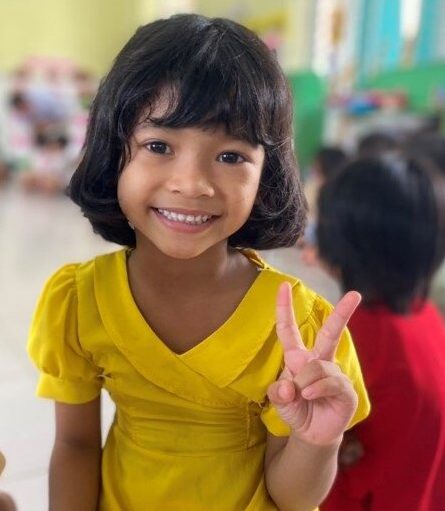 Little girl in yellow dress smiles, holding up peace sign