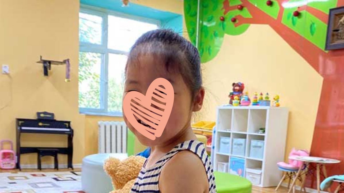 A little girl in a striped dress stands in a brightly decorated room and smiles at the camera