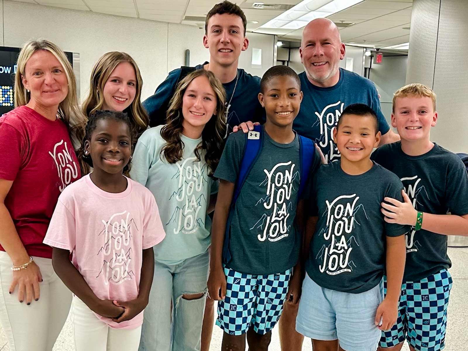 an adoptive family is united with their adopted son at the airport. each family member wears a shirt that says "one day will be the day" in Haitian Creole.